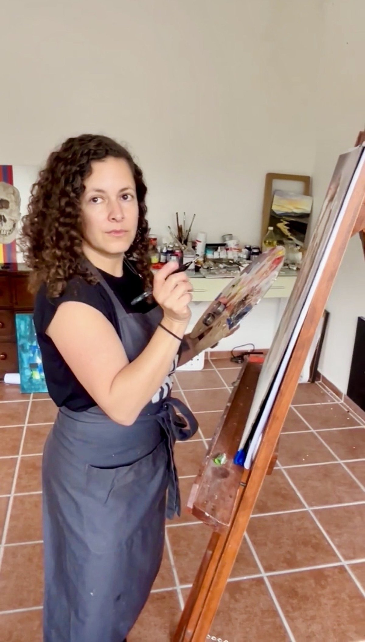 Woman with curly brown hair painting on canvas at an easel in an art studio